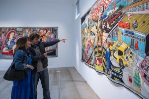 Two people looking on to a huge tapestry in a gallery with clean white walls and another tapestry in the background