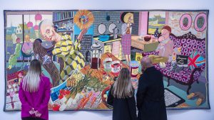 A huge colourful tapestry on the wall depicting various people in a domestic setting. The tapestry is on the wall taking up most of the image, with three bystanders in front looking on