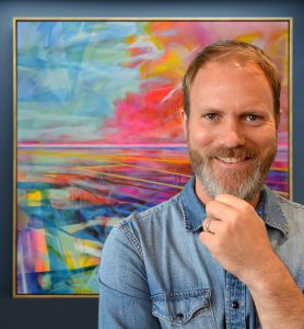 A man, Alex Ebdon, smiles at the camera with his hand touching his chin. He stands in front of a brightly coloured abstract picture.