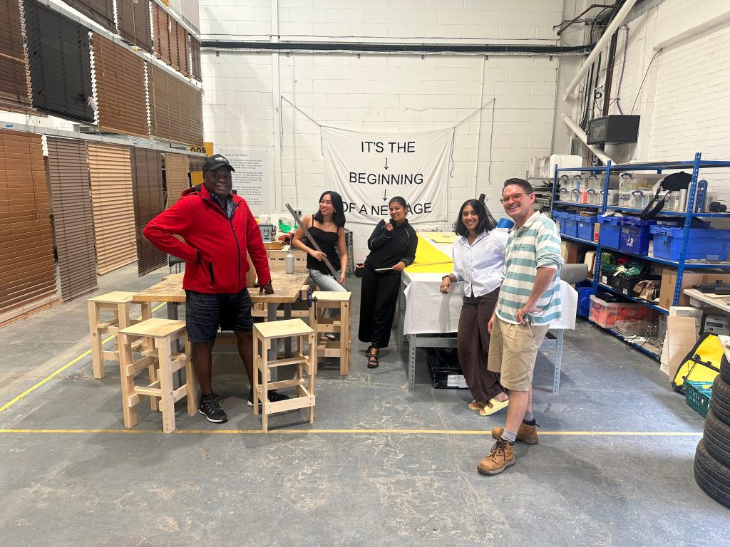 Five people standing in an industrial space with concrete floor and wooden table and chairs.