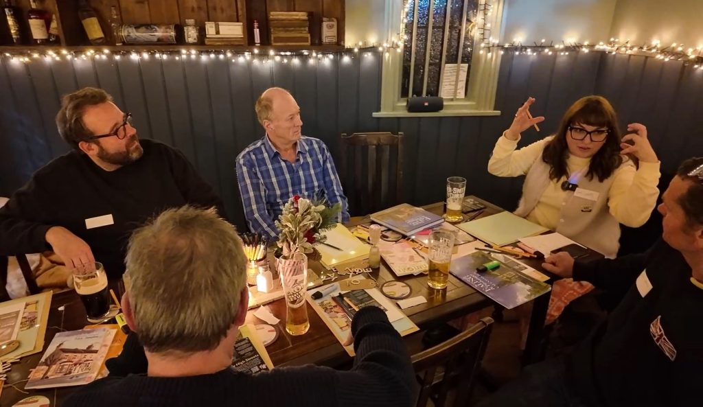 Four men and a woman sitting round a table with pint glasses and stationery in a pub with ambient lighting.