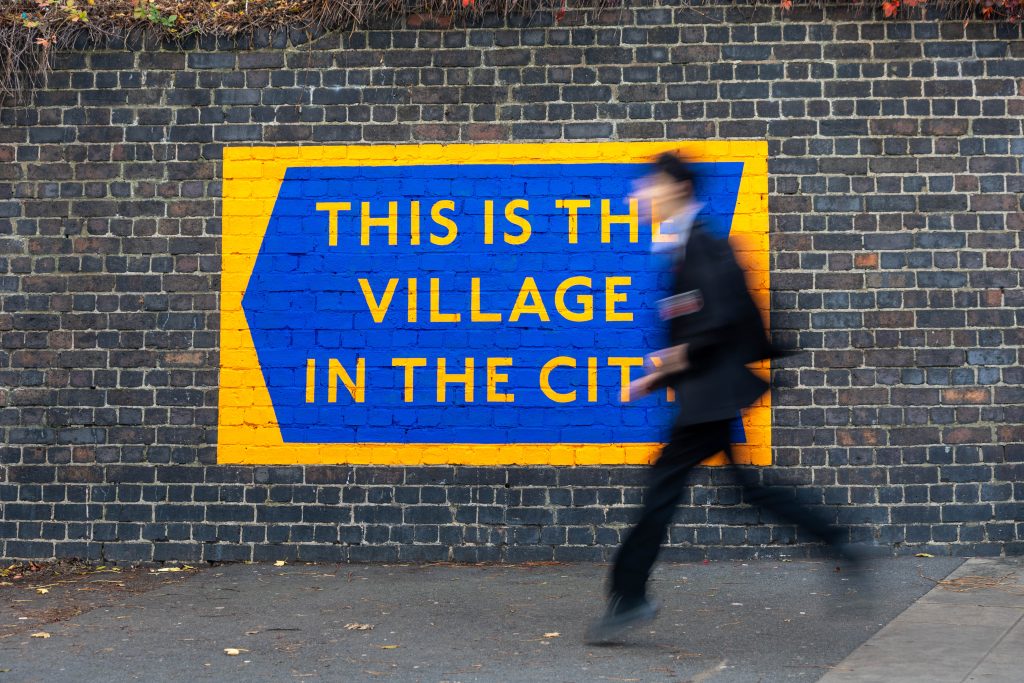 Village in the City (image credit Alex Sturrock) Orange and blue sign on brick wall with blurred figure of schoolboy walking past