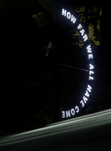 How Far We All Have Come (image credit Alex Sturrock) Illuminated sign, white lettering in a semi-circle, black night, streak of white beneath.