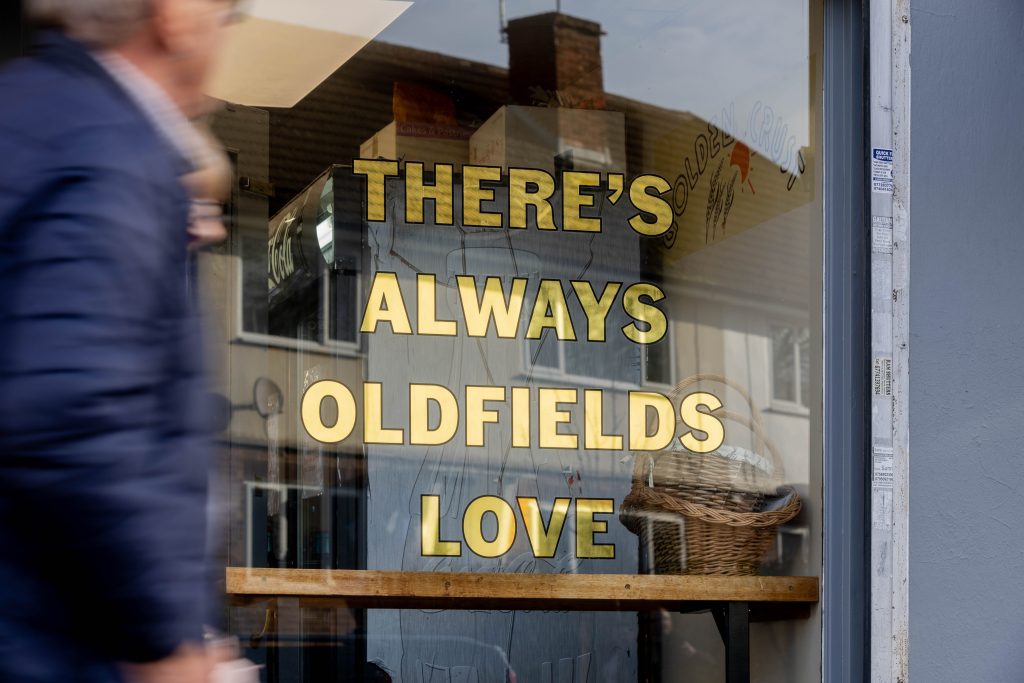 Oldfields Love (image credit Alex Sturrock) Gilded lettering in shop window. House reflected in glass. Blurred figure of man in navy coat walking past.