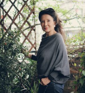 A woman in a baggy grey top stands outside with greenery and a trellis behind her.
