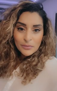 A woman with long blond curly hair in close up looking straight at the camera, wearing.a beige top