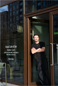 A man wearing black with his arms folded leans against the door frame of a cafe.