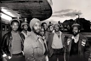 A black and white photo of seven people stood under an underpass looking at the camera, wearing 70s style clothing - this is Misty in Roots.
