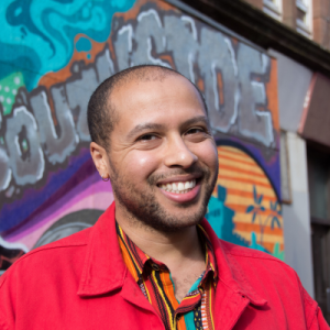 A man with short dark hair smiling at the camera. He is wearing a red shirt and stands in front of a wall with graffiti on.