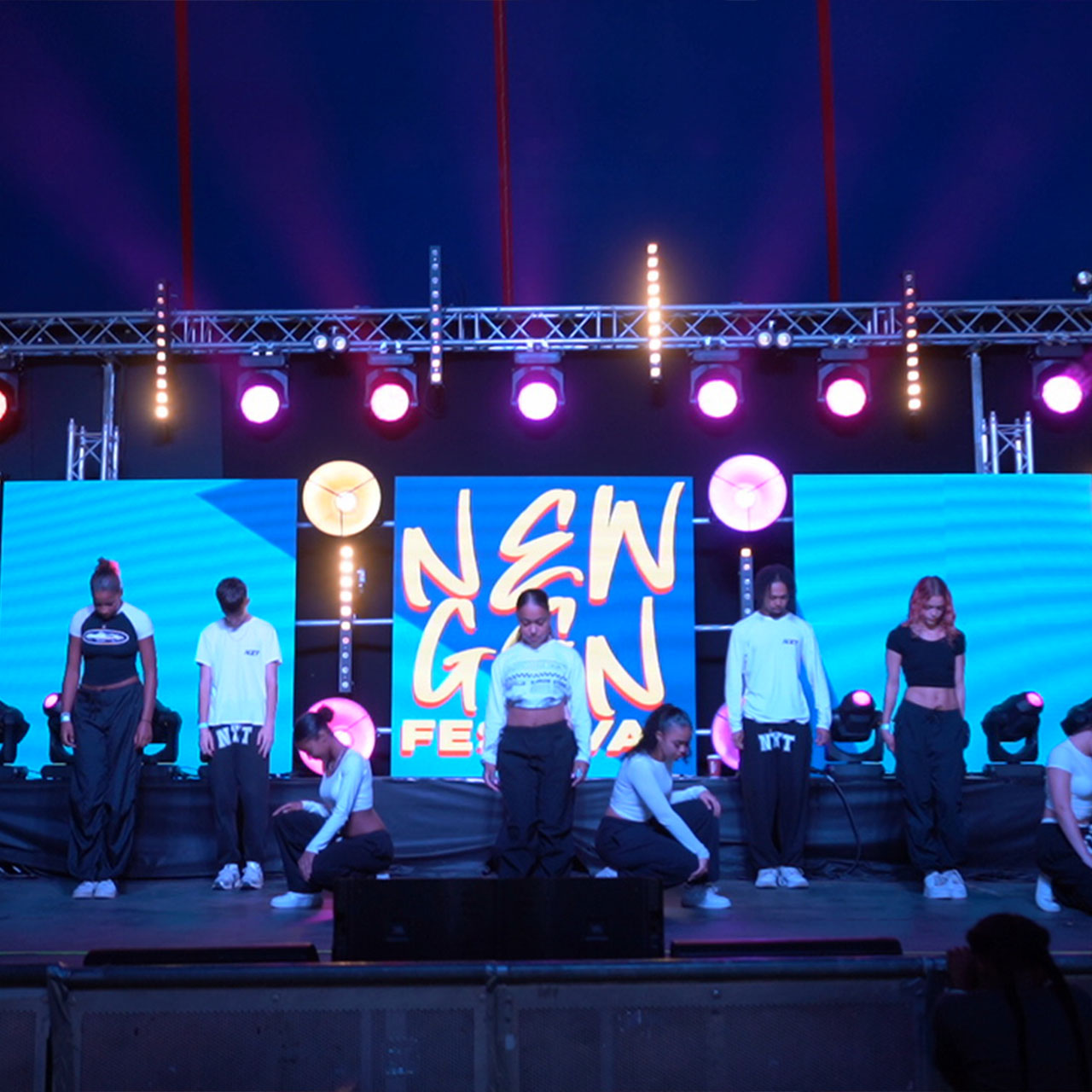 A stage with blue and orange lighting and 'New Gen Festival' in orange writing on a blue background. There are a few performers on the stage