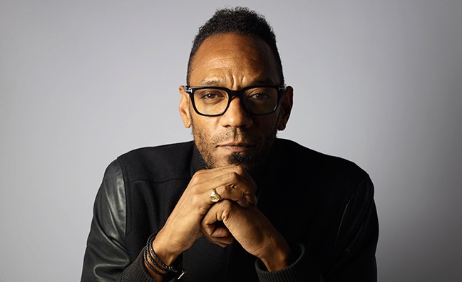 A man in a black shirt and glasses rests his head on his hands and looks into the camera. Against a white-grey background.