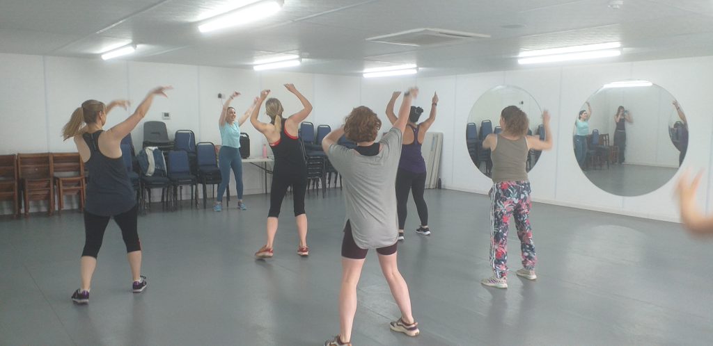 Six people dance in a room with circular mirrors on the wall, in front of them an instructor leads them