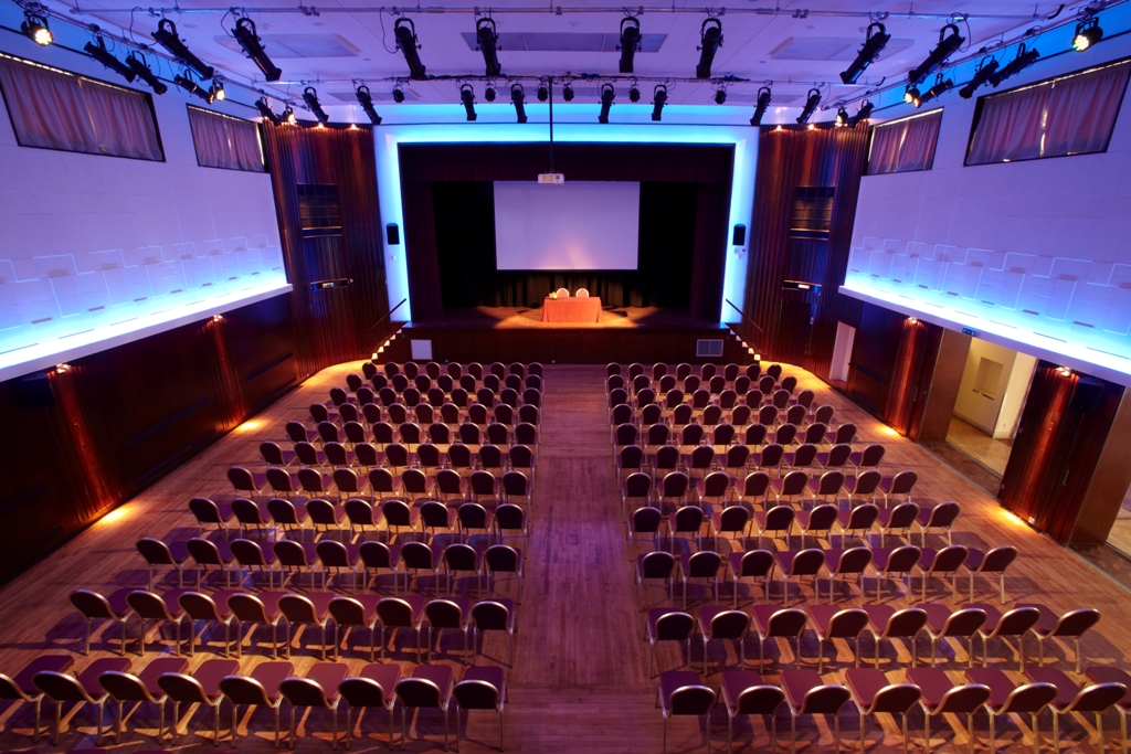 A large room with wooden floorboards and black chairs arranged conference style seen from the back of the room, There is blue and orange lighting and a screen lit up on a stage at the far end of the room