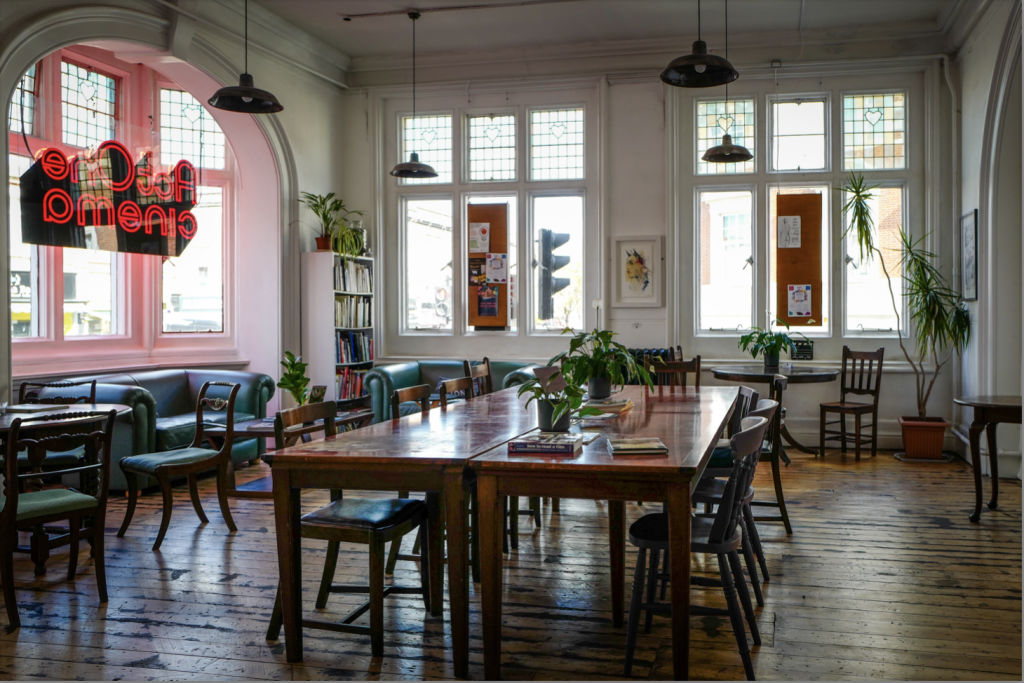 A room with about ten dark wooden chairs around a dark wooden table with two house plants on. Around the medium sized room there are two turquoise leather sofas, more wooden chairs and large windows. A red light in the shape of 'ActOne cinema' hangs in the window to the left.