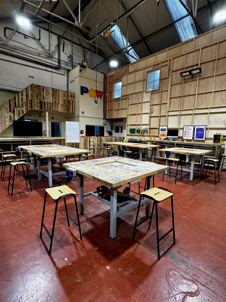 A large warehouse with wooden tables and stools on a rough red floor