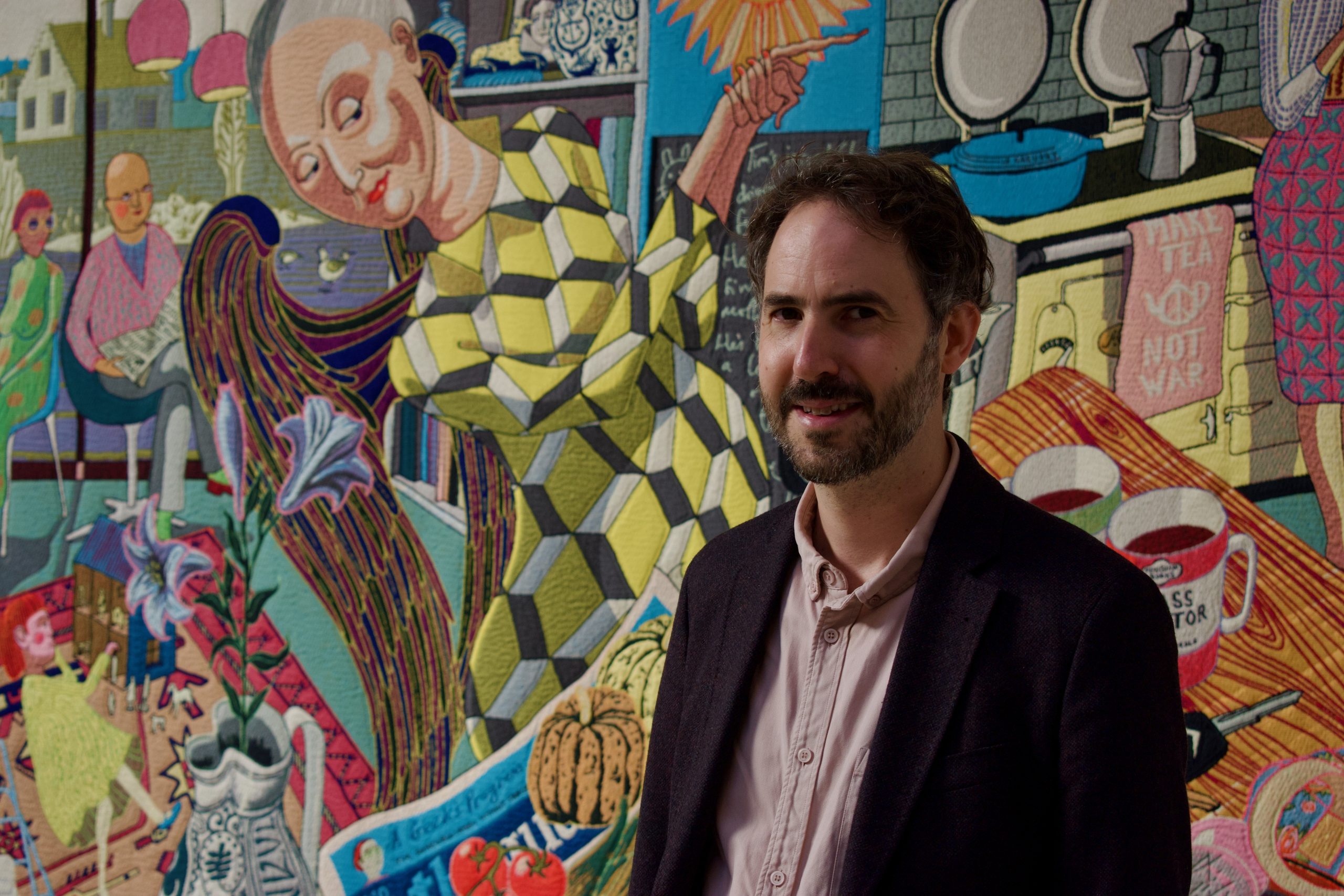 A portrait of a man in a shirt and dark jacket, smiling at the camera in front of a large colourful tapestry depicting people in a domestic setting