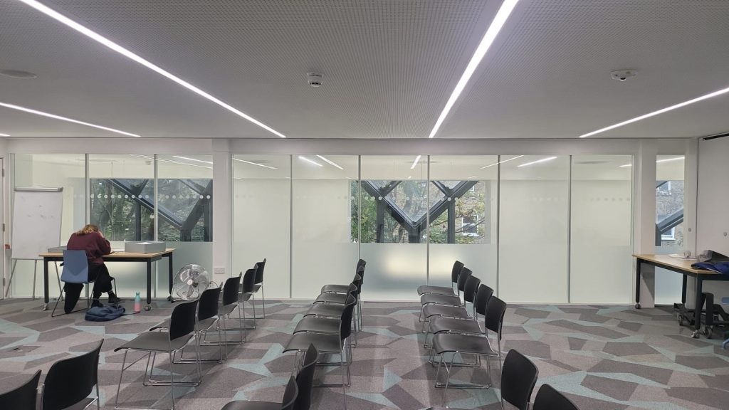 Rows of black chairs in an almost empty room. There is someone sat at a table in the background