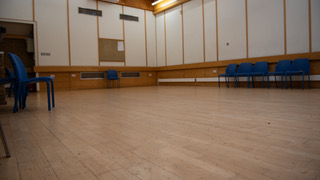 A large empty room with wooden floorboards and white walls. A few blue chairs are scattered around the outside of the room.