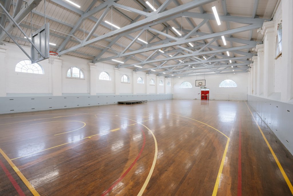 Sports hall with wooden floor, basketball hoops, steel rafters, and half-moon windows
