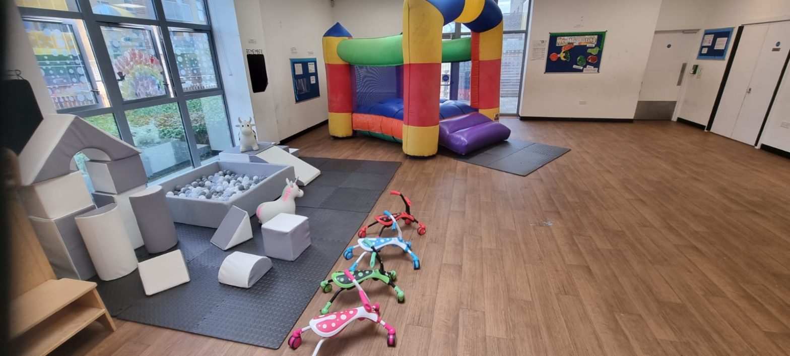 A large room with wooden flooring, white walls, large windows. There is a bouncy castle in the background and a selection of foam shapes and toys in the foreground.