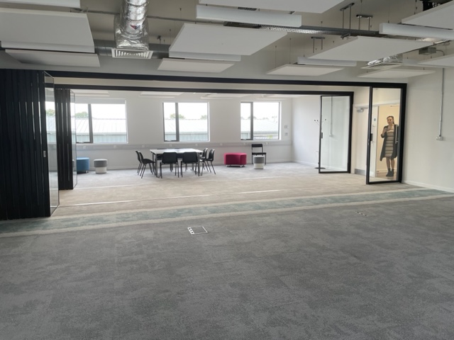 A large space with a table in the background with chairs around it, grey flooring and a person stands in the doorway on the right