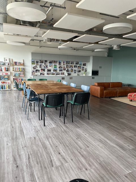 A modern space with grey floorboards, bookshelves and pictures on the wall at the back, a large brown table in the middle with green chairs around it and a brown leather seating area on the right