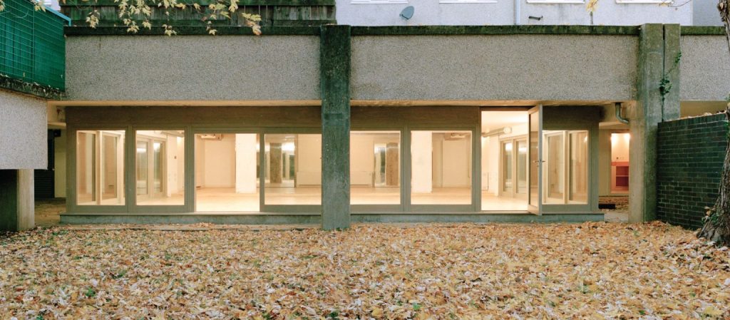 A white building with windows all along the front set in a garden with yellow and green on the floor - it's indistinguishable but may be autumn leaves filling the ground.
