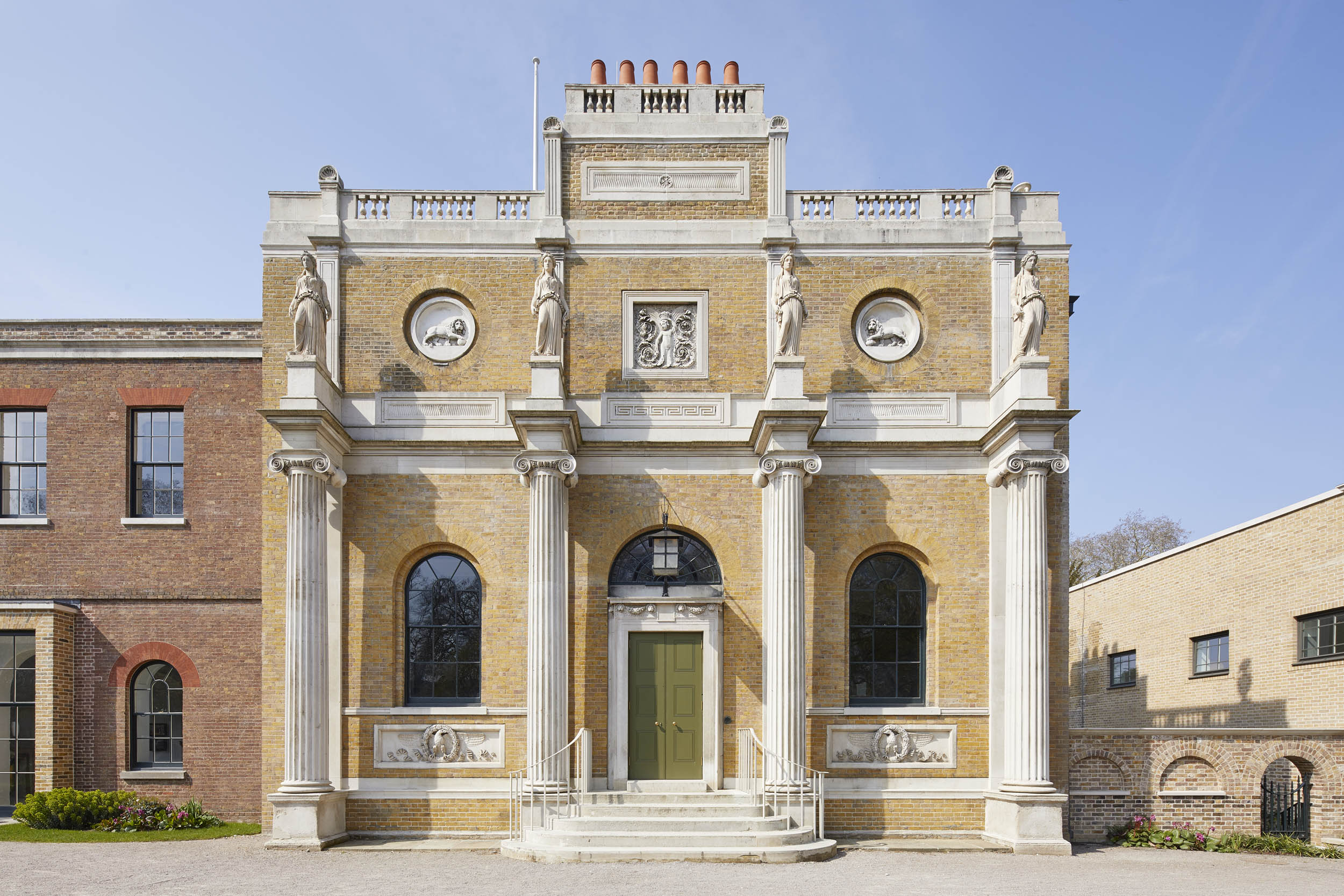 A grand bright Manor House with four white pillars on the front, back with a white statue stood on them on a sunny day with a blue sky and no clouds