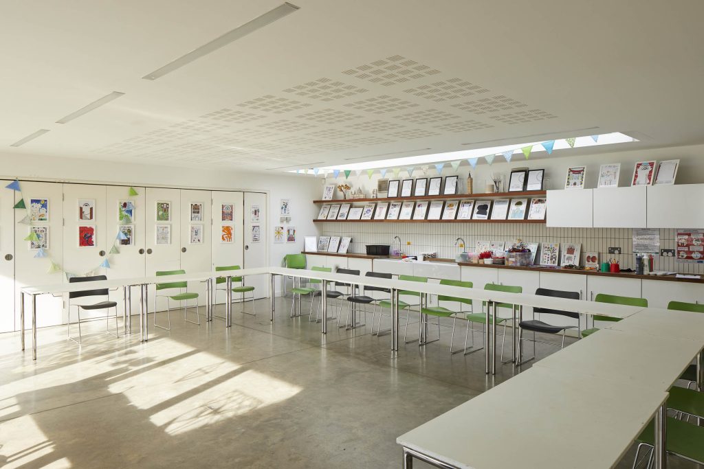 An empty, modern classroom, with long white tables in a half circle, green and black chairs around the tables and lots of small framed pictures on the wall