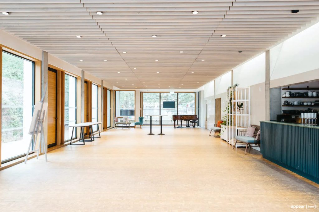 A large, empty room with huge windows lighting the room with daylight. There is a green bar to the right of the room and a few chairs and small tables in the room