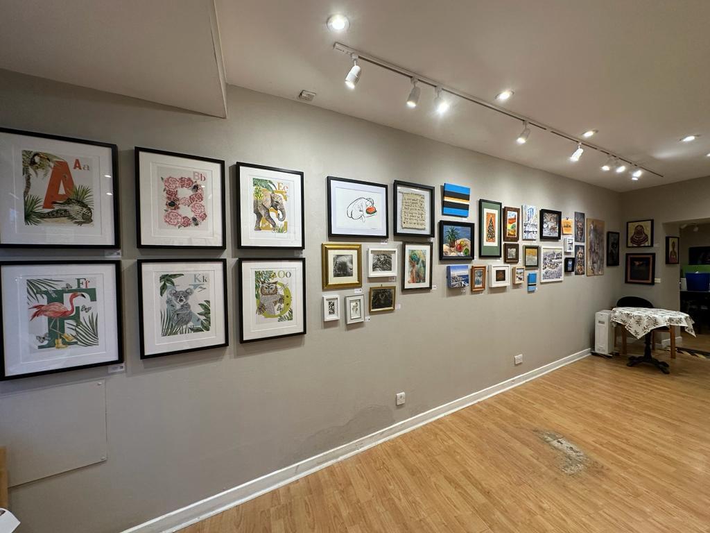 The side of an empty room with lots of modern artworks in frames o the wall. Wooden floorboards and a small table in the background with a white cloth on.