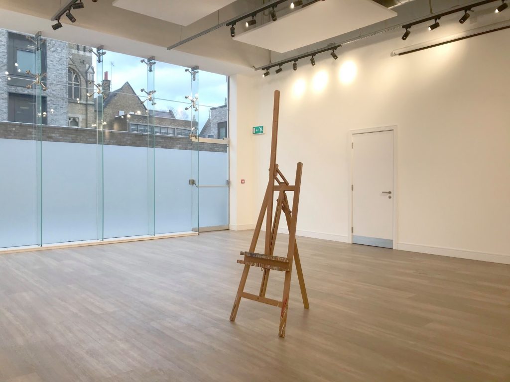 A studio space with a wooden easel placed in the middle, cream walls on the right and a window on the left with old buildings behind