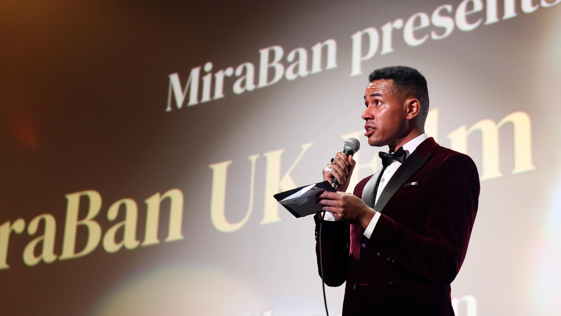 A man holds a microphone looking out ahead and in his left hand holds an envelope. Behind him, a large screen has a logo for MiraBan UK Film