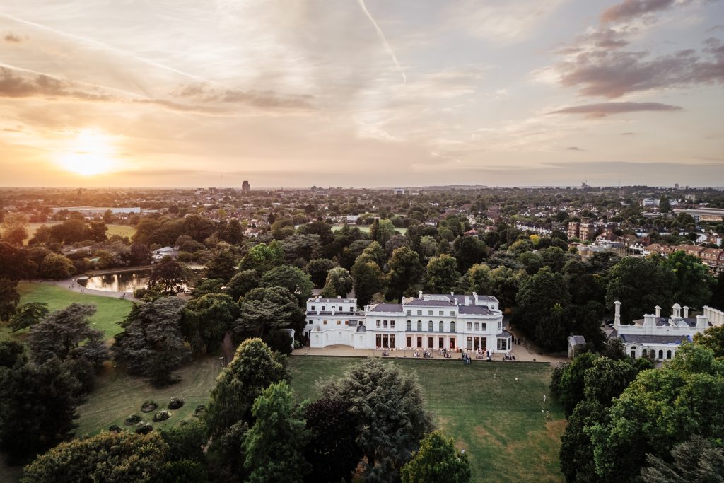 A grand Manor House set in huge gardens with large, old, green trees surrounding it. The sun is setting in the background and clouds are scattered across the dimming sky