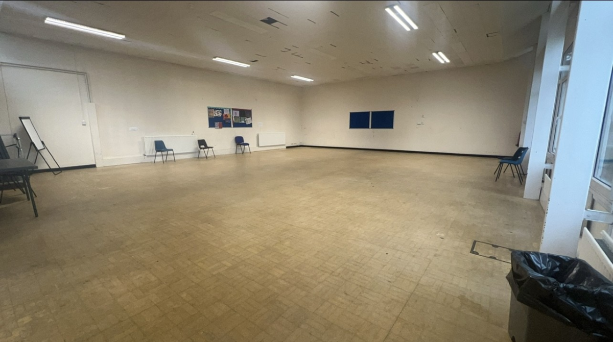A large open room with a four black chairs on the outside of the room. Wooden tiled floor and florescent lights on the ceiling.