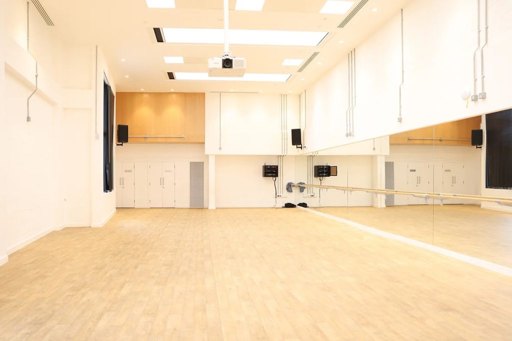 An empty room with no furniture. Wooden floor boards, wooden panels on the wall, mixed with grey walls, an internal window in the background and ballet barre attached to a mirrored wall on the right