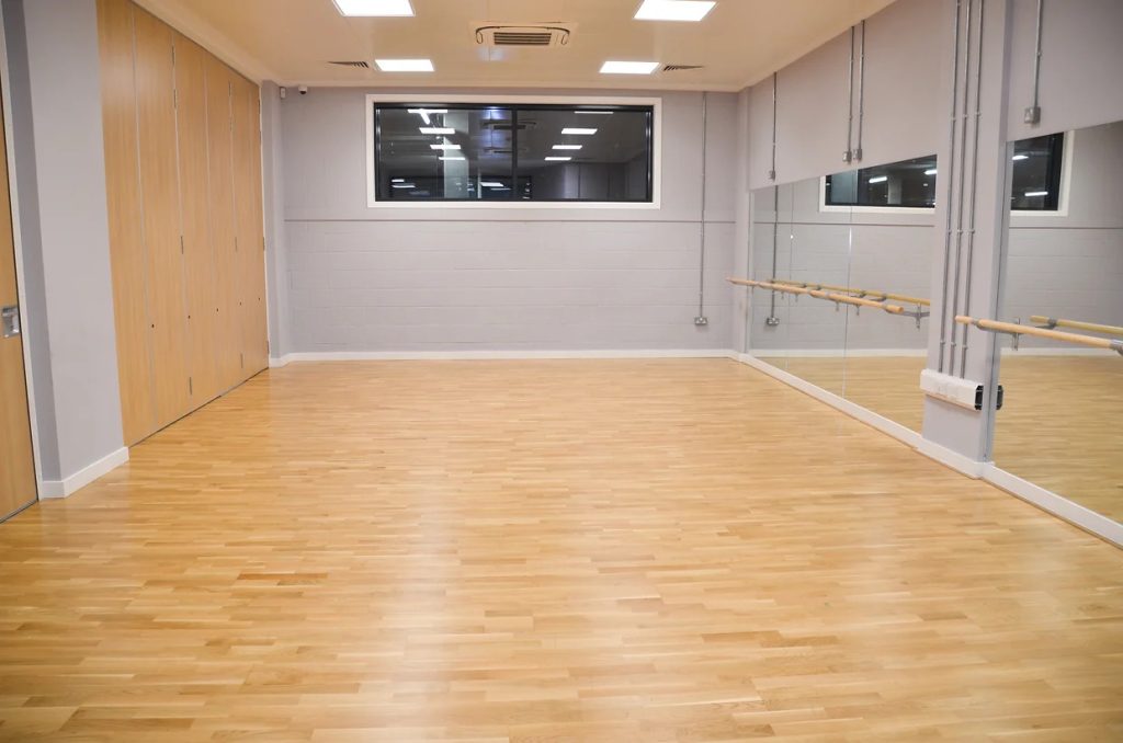 An empty room with no furniture. Wooden floor boards, wooden panels on the wall, mixed with grey walls, an internal window in the background and ballet barre attached to a mirrored wall on the right