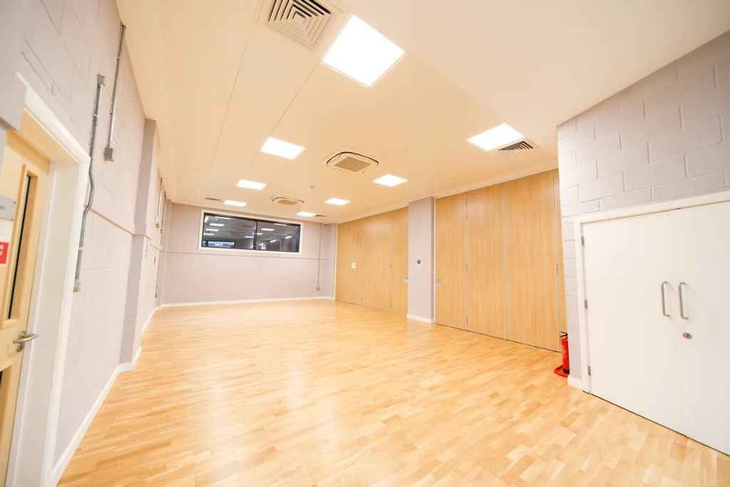 An open room with no furniture. Wooden floorboards, a door on the left, an internal window in the background and white cupboard on the right.