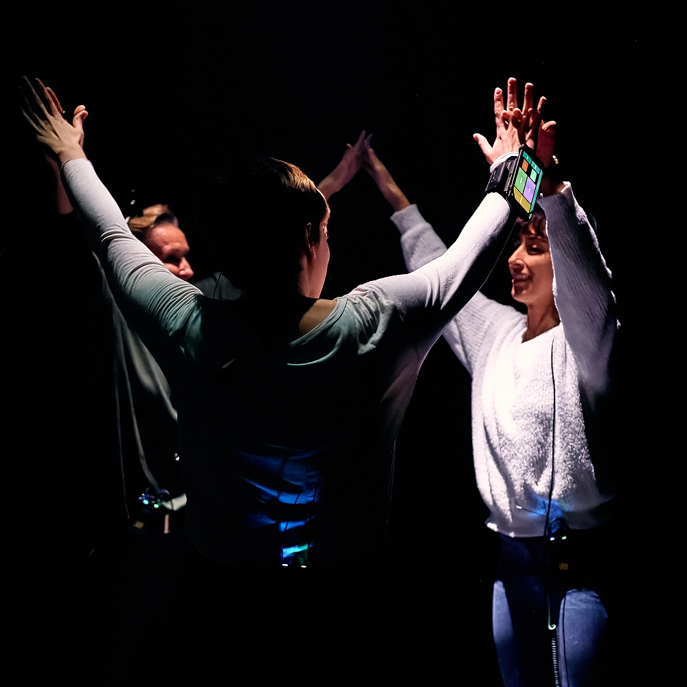 Three people stand in a circle with their arms raised in the air in a dark, black space