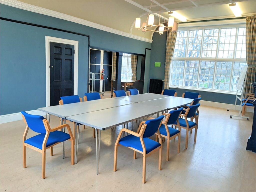 Ten blue chairs around four white tables put together to make on big table. Light flooring, a large window in the background, blue walls and a black door on the left