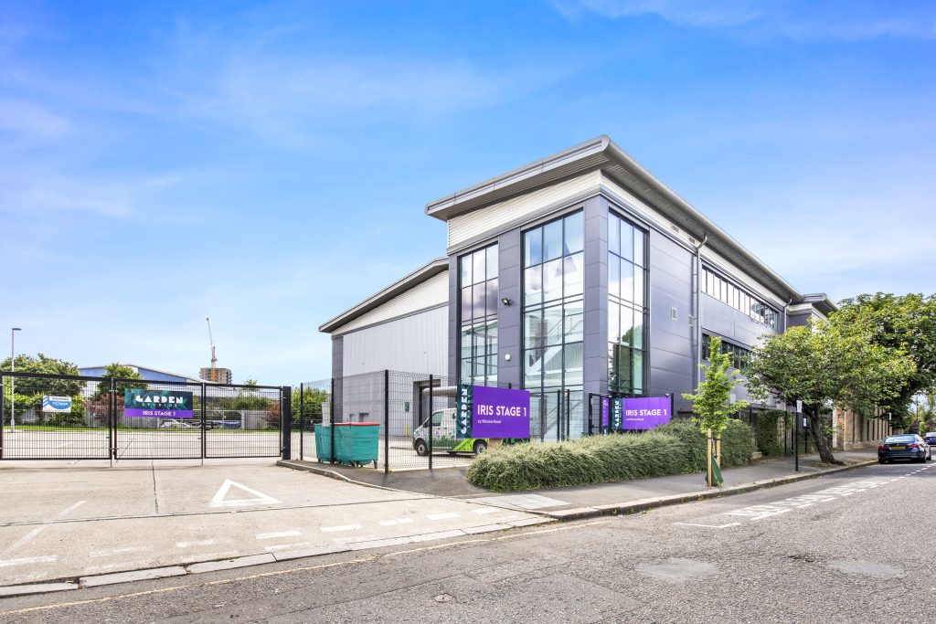 A very modern building with big windows sat inside a gated area with road around it on a sunny day