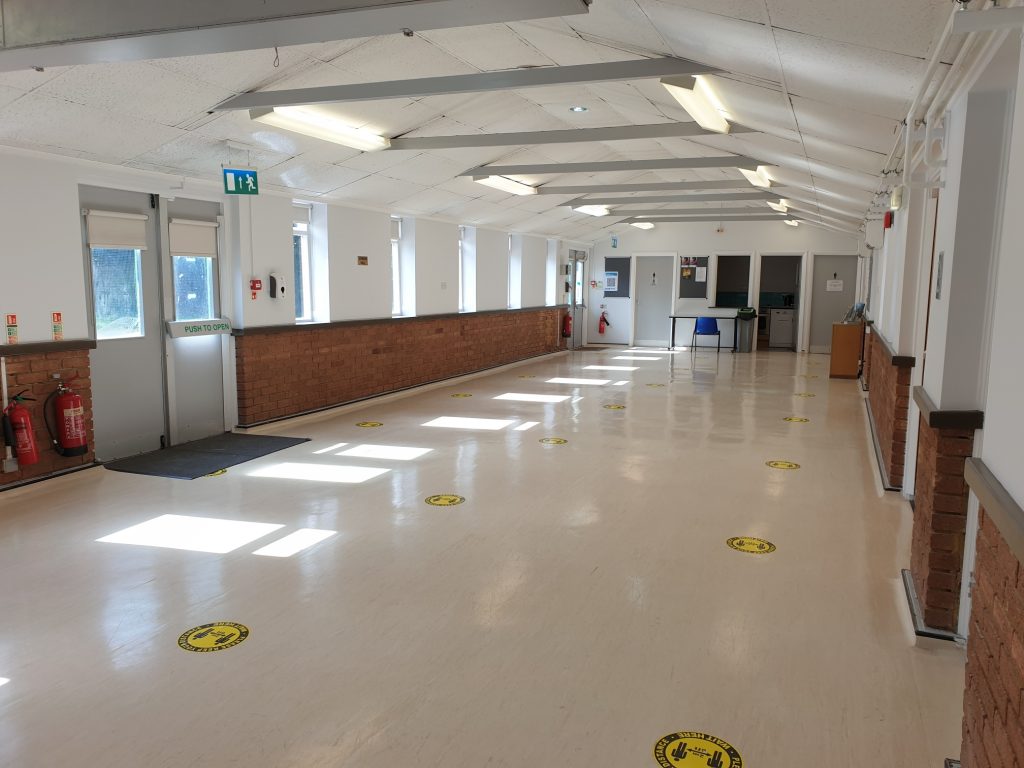 A long, large, empty room with a blue chair in the far distance. The floor is shiny and light white-brown colour, the walls are red brick to about a metre and then white above, white ceiling and a row of several windows on the left