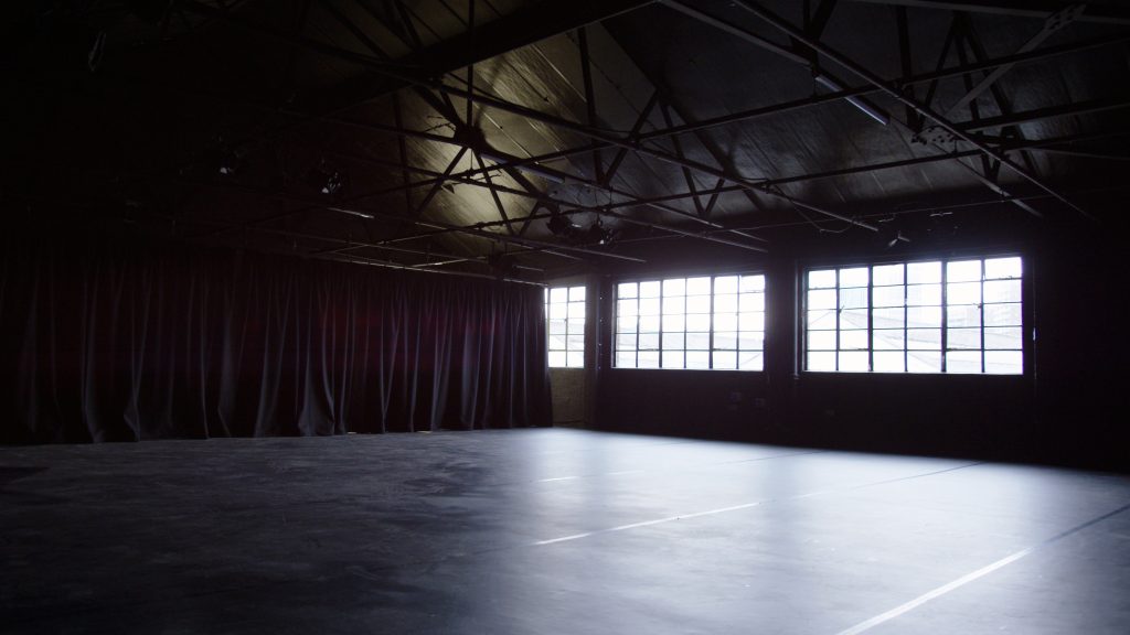 A large dark room with dark floor, dark ceiling with rigging on, a dark curtain pulled halfway around the room on the left and windows on the right