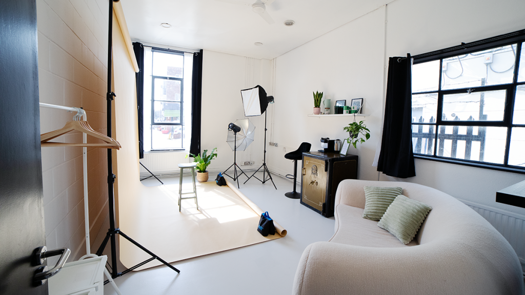 A photography studio with an infinity wall on the left, a cream sofa on the right, lights, tripods and camera equipment in the background, with white walls and floor and two large windows with black curtains.