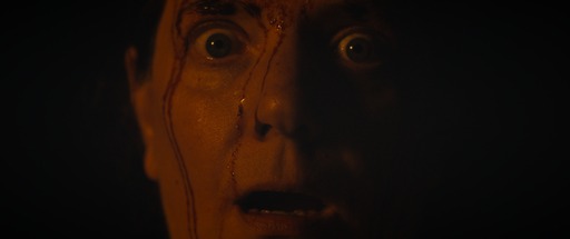 Extreme close up of a woman's face with wide eyes and open mouth, three lines of blood fall down her face. It is a dark image with low lighting and a black background