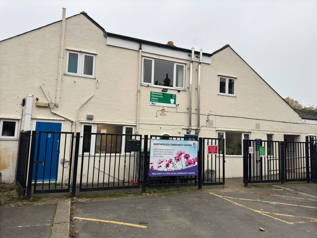 A cream building with two peaks in the roof, a carpark in front and a sign saying 'Northfields Community Centre'