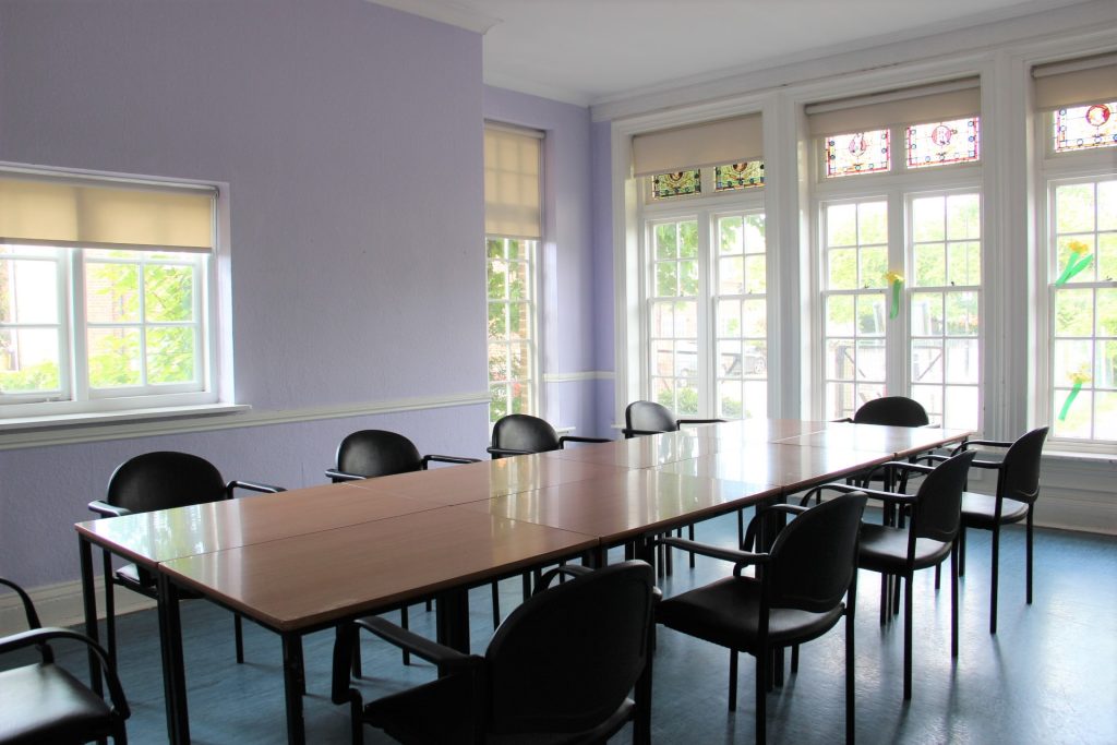 Ten black chairs around a conference table in a room with blue flooring, light purple walls and many windows