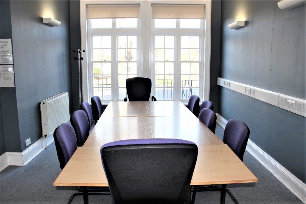 Ten purple chairs around a light brown table in a small room with grey carpet, dark grey walls and a window at the back