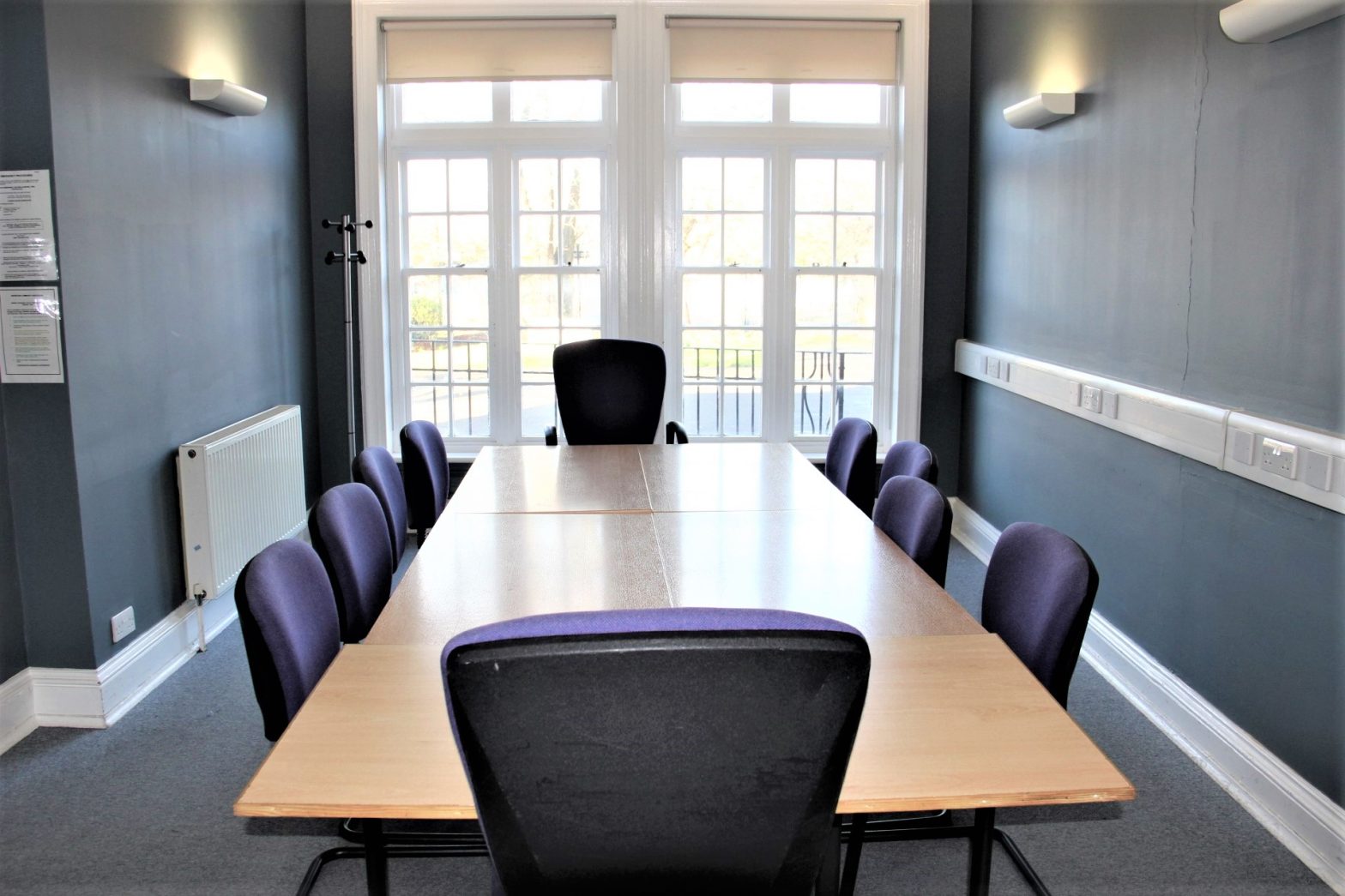 Ten purple chairs around a light brown table in a small room with grey carpet, dark grey walls and a window at the back