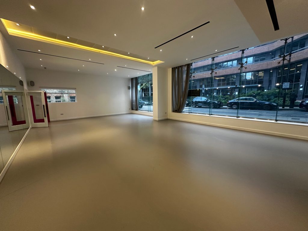 A large empty studio with light brown flooring, a mirror wall on the left and a large window looking out to parked cars in the road on the right
